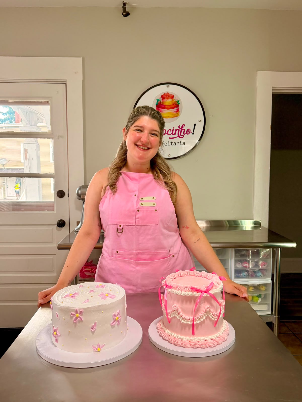 Paula sorrindo com avental rosa ao lado dos bolos que decorou